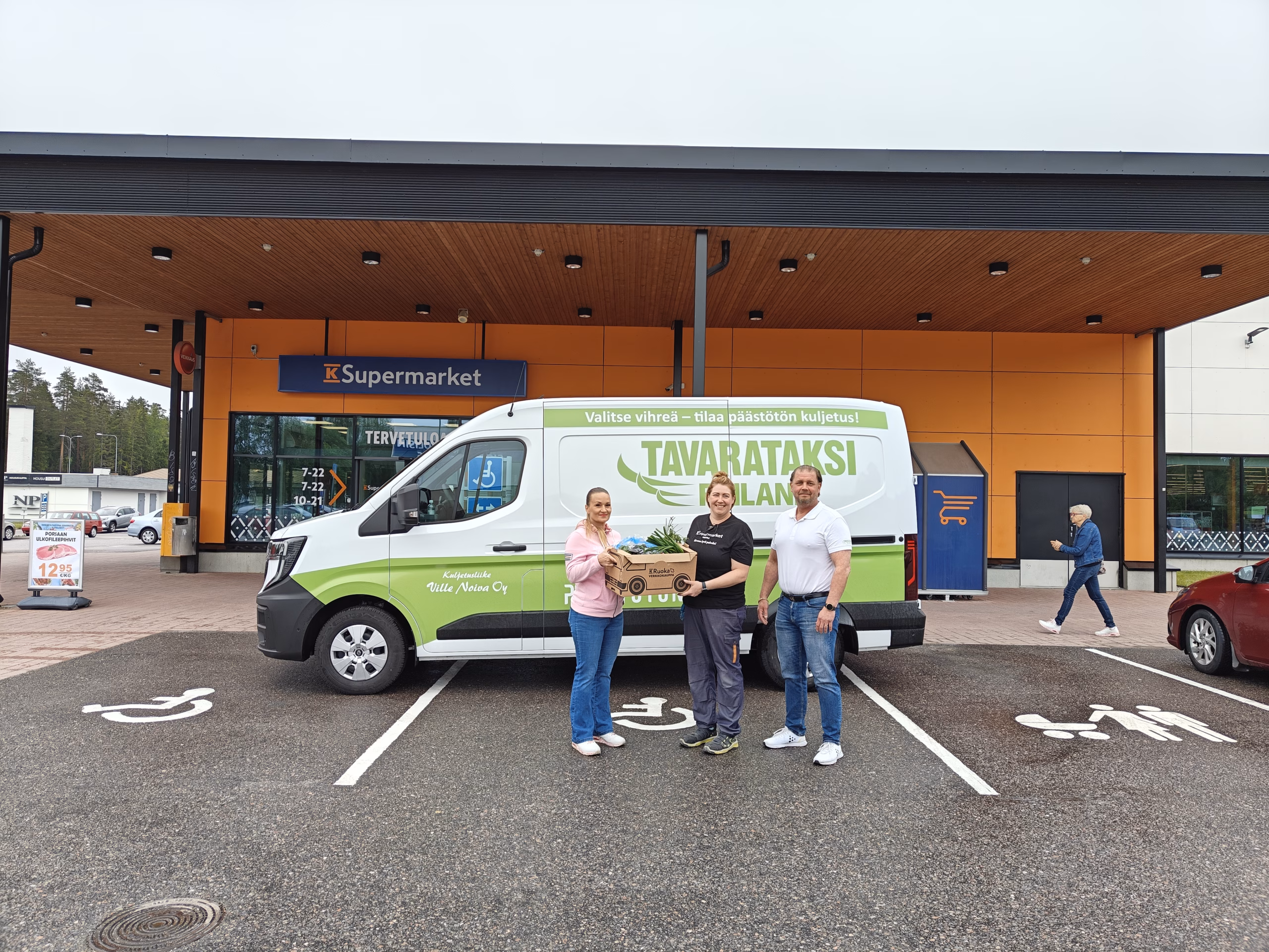 KUvassa K-Supermarket-kauppiaita sekä Tavarataksin edustaja ja Tavrataksin sähköinen jakeluauto. Kaupan edustalla poseeraten.